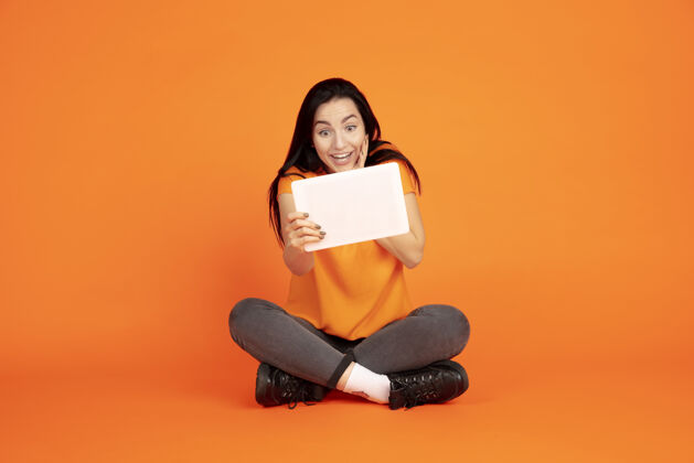 员工橙色工作室背景上的白种人年轻女子肖像穿着衬衫的漂亮黑发女性模特人类情感的概念 面部表情 销售 广告文字空间使用平板电脑 虚拟博客面部女商人工作