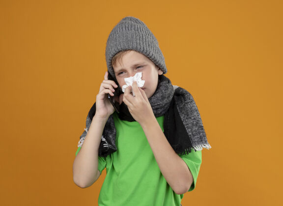 感冒生病的小男孩穿着绿色t恤 戴着暖和的围巾 戴着帽子 感觉不舒服 拿着手机说话 用纸巾擦鼻子 站在橙色的墙上受冻餐巾小鼻子