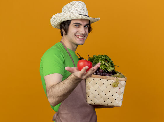 西红柿年轻的园丁 围着围裙 戴着帽子 手里拿着装满蔬菜的箱子 站在橙色的墙上 看到新鲜的西红柿笑着 脸上洋溢着幸福围裙板条箱脸
