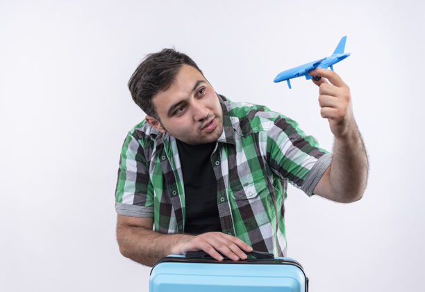 假期穿着格子衬衫的年轻旅行家拿着手提箱站着 手里拿着玩具飞机 脸上挂着幸福的笑容 越过白墙旅行脸衬衫