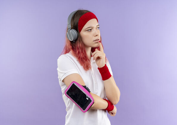 人头戴耳机 脸上带着沉思表情 戴着智能手机袖标站在紫色墙上的年轻运动女性人表情运动装