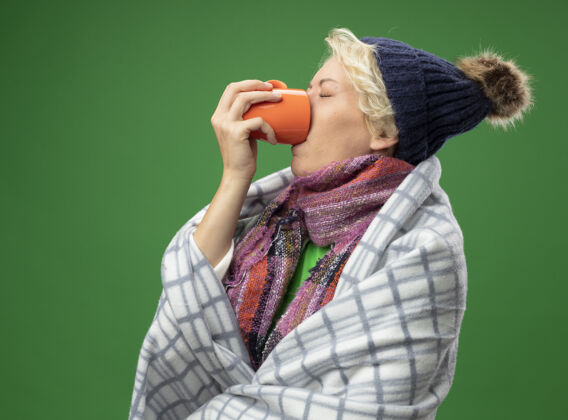 包裹生病的不健康的女人 短头发 戴着暖和的围巾和帽子 感觉不舒服 裹着毯子 喝着热茶站在绿色的墙上头发不健康喝酒