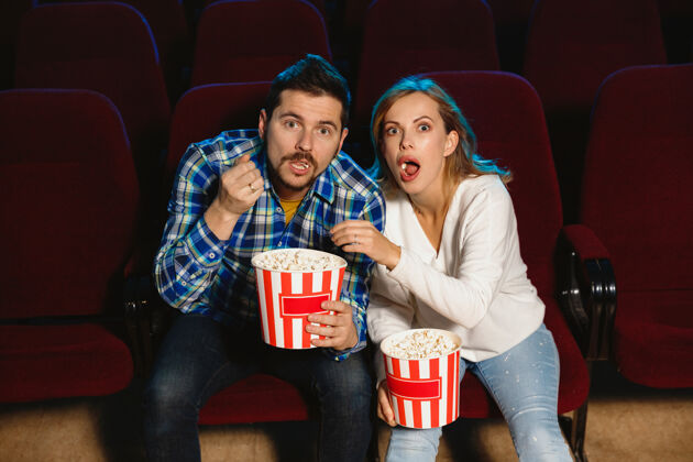 吃迷人的年轻白种人夫妇在电影院 房子或电影院看电影看起来富于表现力 惊讶和情绪化独自坐着玩关系 爱情 家庭 周末时间椅子表达金发