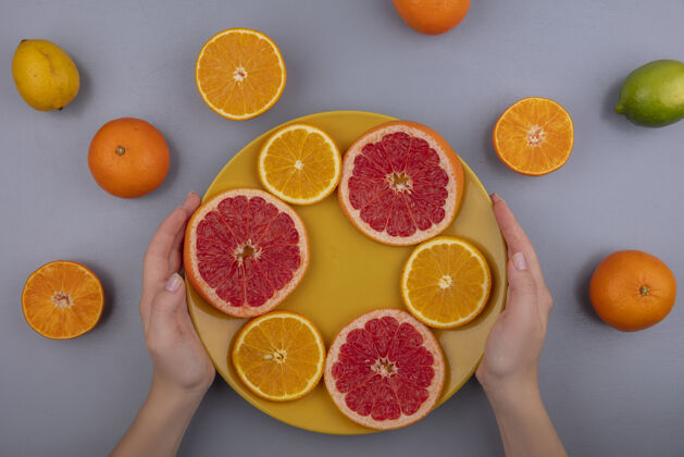 视野俯视图：灰色背景下 黄色盘子上拿着葡萄柚片和橘子的女人切片新鲜葡萄柚