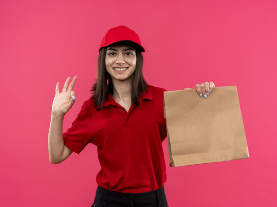 年轻年轻的送货员身穿红色马球衫 戴着帽子 手里拿着纸包 面带微笑 粉色的墙上挂着ok标志展示看包装