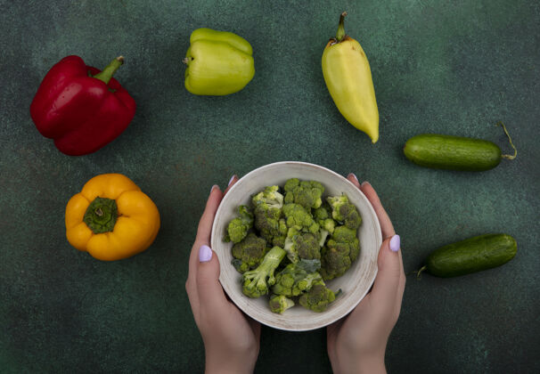 食物俯视图：一个女人手里拿着一碗西兰花 绿色背景上有黄瓜和甜椒碗绿色顶部