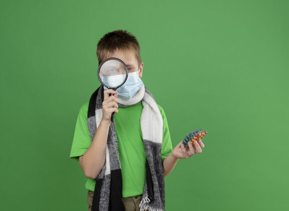 放大镜生病的小男孩穿着绿色t恤 脖子上围着暖和的围巾 戴着护面面具 手里拿着药片 站在绿色的墙上透过放大镜看抱着面部小