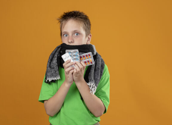 疾病生病的小男孩穿着绿色t恤 脖子和嘴上围着温暖的围巾 站在橘色的墙上 他看起来很担心看围巾小