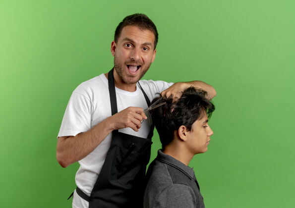 客户快乐的理发师围着围裙剪头发 满意的顾客站在绿色的墙上帅哥头发站