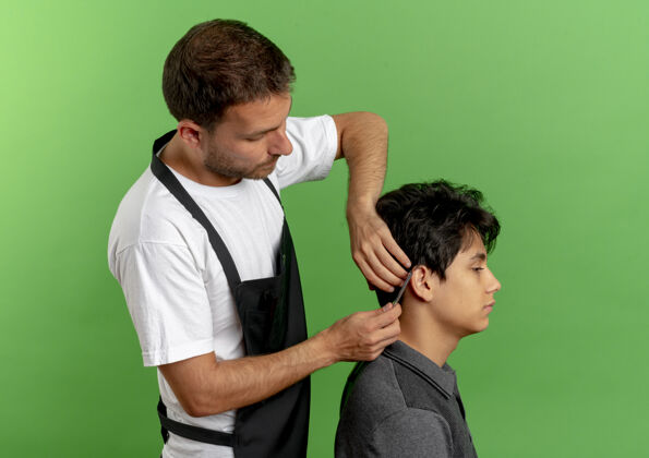 工人穿着围裙的理发师用剪刀剪头发 满意的顾客站在绿色的墙上工作剪刀男人