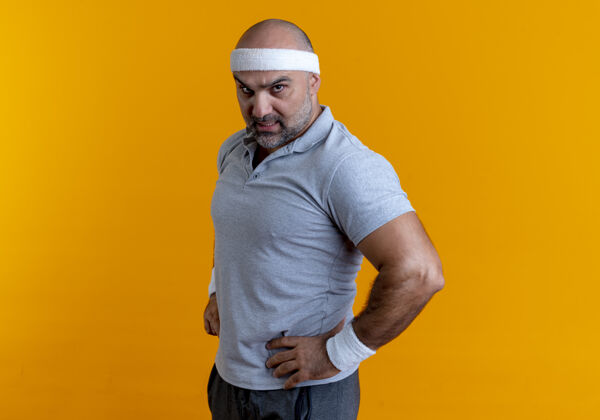运动员戴着头巾的成熟的运动型男人向前看 胳膊放在臀部 严肃的脸站在橙色的墙上运动运动装成熟