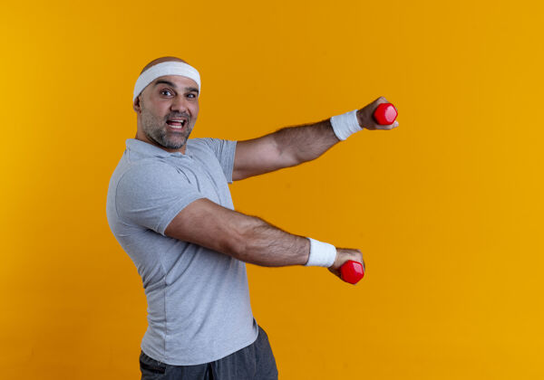 人成熟的运动型男子戴着头巾 手持哑铃 站在橙色的墙上 自信地微笑着工作头带男人