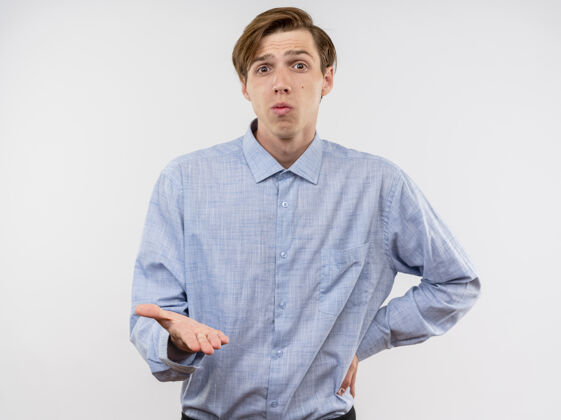 男人一个穿着蓝色衬衫 伸出胳膊的年轻人站在白色的墙上感到困惑和不快年轻看着手臂