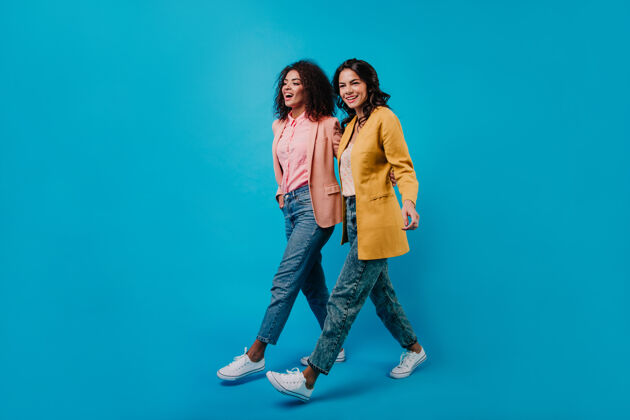 女性两个时髦女人在蓝色墙壁上行走的全长摄影棚镜头时尚镜头年轻