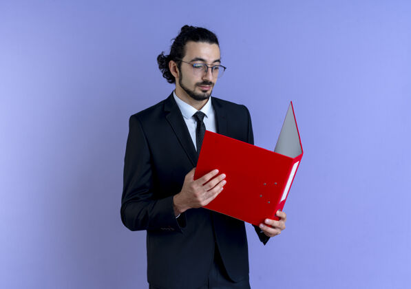 男人一个穿着黑西装 戴着眼镜 拿着红色文件夹的商人站在蓝色的墙上严肃地看着它西装脸男人