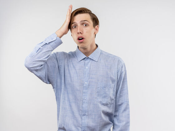 困惑穿蓝衬衫的年轻人正困惑和担心 手放在头上 因为他站在白墙上犯了错误看年轻站