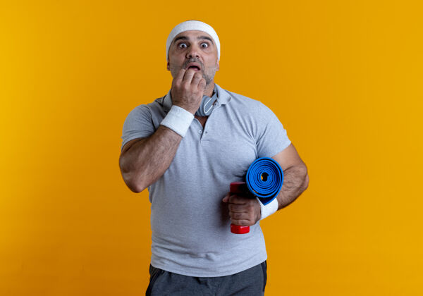垫子成熟的运动型男人戴着头巾 拿着瑜伽垫 站在橘色的墙上 看上去既惊讶又惊奇人人男