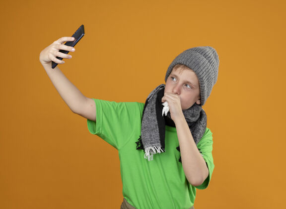少生病的小男孩穿着绿色t恤 戴着暖和的围巾和帽子 感觉不舒服拿着智能手机自拍 站在橙色的墙上咳嗽自拍男孩疾病