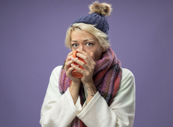 温暖生病的不健康的女人 短头发 戴着暖和的围巾和帽子 感觉不舒服 站在紫色的墙上喝着热茶站不适生病