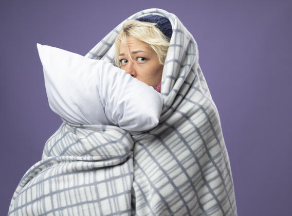 悲伤生病的不健康的女人 短发 戴着暖和的帽子 裹着毯子 抱着枕头 不舒服 悲伤的表情 担心紫色的墙壁表情短裤头发