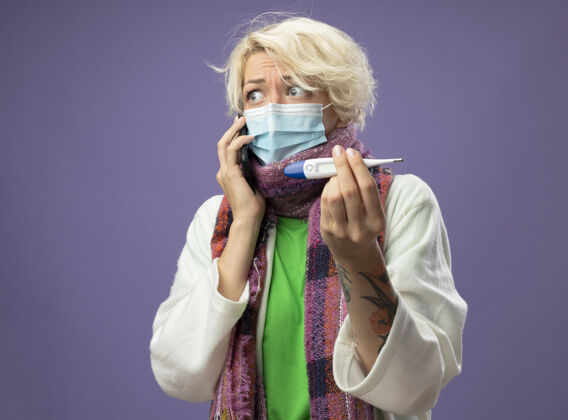 围巾生病的不健康的女人 短头发 戴着暖和的围巾 戴着面罩 拿着温度计 站在紫色的墙上讲手机 看起来很担心抱着说话体温计