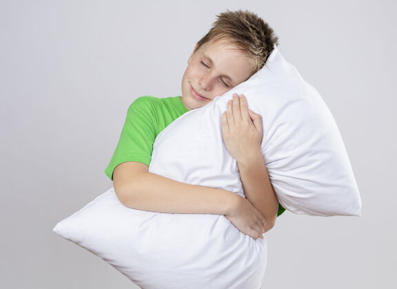 枕头生病的小男孩穿着绿色t恤抱着枕头 闭着眼睛微笑着站在白色的墙上闭上小站立