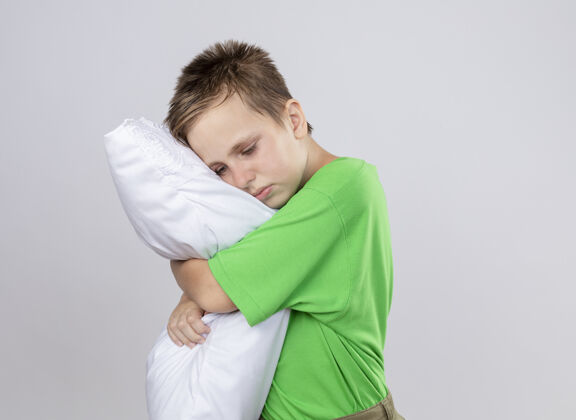 疾病生病的小男孩穿着绿色t恤感觉不舒服抱枕闭着眼睛站在白色的墙上不适枕头小