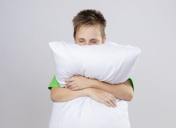站立生病的小男孩穿着绿色t恤感觉不舒服抱枕闭着眼睛站在白色的墙上不适感觉枕头
