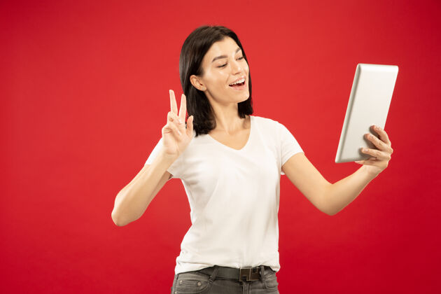 行政红色工作室背景上的白人年轻女子半身像穿着白衬衫的漂亮女模特人类情感的概念 面部表情使用平板电脑制作视频博客或聊天年轻黑发手势