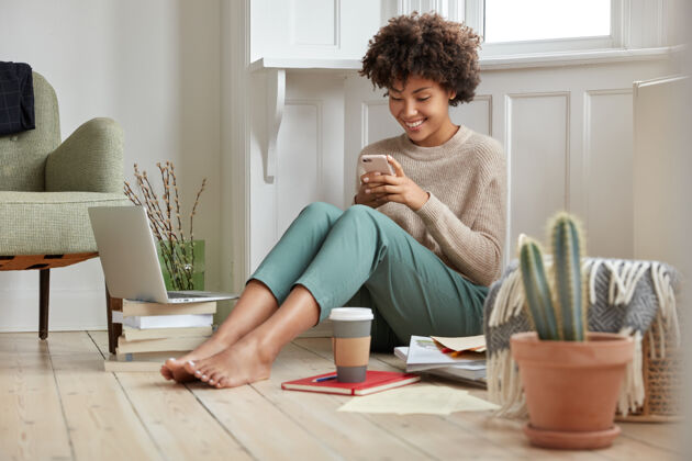 微笑积极的黑人女士喜欢喝咖啡去 手里拿着智能手机 在社交网络中阅读短信 对现代科技和wifi连接感到满意 在家里学习 做研究阅读咖啡电子