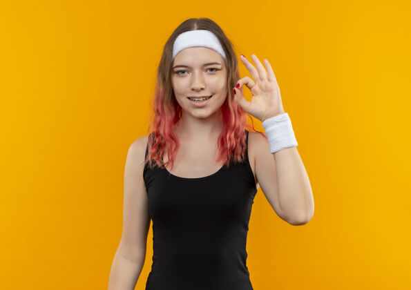 运动装身着运动服的年轻健身女士站在橙色的墙上 愉快地微笑着 展示着ok标志运动员看人