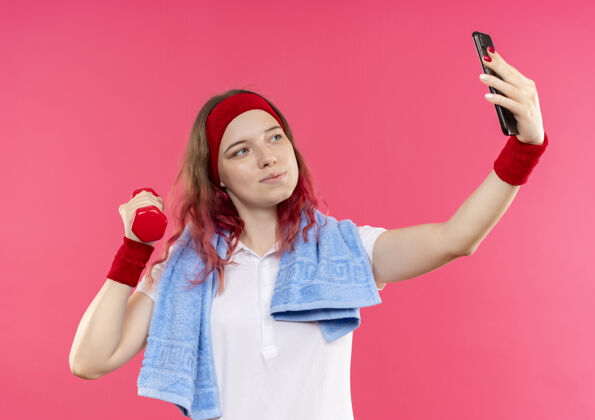 移动戴着头巾 肩上扛着毛巾的年轻运动型女子自拍 手拿哑铃对着站在粉色墙上的智能手机镜头运动姿势肩膀