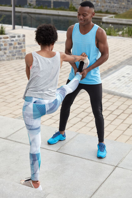 耐力男教练的垂直镜头帮助他的黑人女学员做伸展运动 站在户外运动型女子站在后面 表现出良好的灵活性 抬腿高 穿运动鞋实习生肌肉姿势