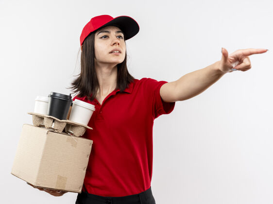 帽子年轻的送货员穿着红色制服 戴着帽子 手里拿着盒装和咖啡杯 一边看一边用食指指着白色背景上的东西拿着年轻人指着
