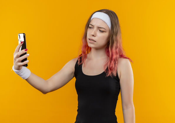 自拍穿着运动服的年轻健身女士用智能手机自拍 严肃的脸站在橙色的墙上采取市民年轻