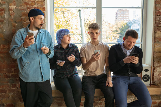 内容一群快乐的白人年轻人站在窗户后面分享新闻 照片或智能手机上的视频 聊天或玩游戏 享受乐趣社交媒体 现代科技移动计算机无线