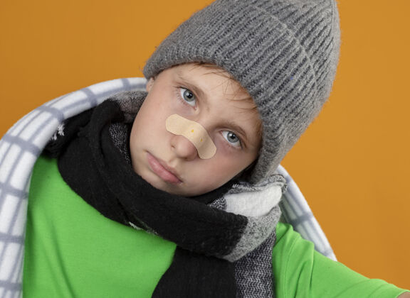 补丁生病的小男孩戴着暖和的帽子 戴着围巾 裹着毯子 鼻子上贴着补丁 看着站在橙色背景下的摄像机 他既不高兴又不舒服不高兴包生病