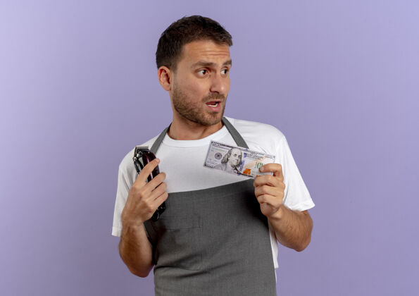 男人围裙里的理发师手里拿着理发机和现金站在紫色的墙上困惑地看着旁边看工作头发