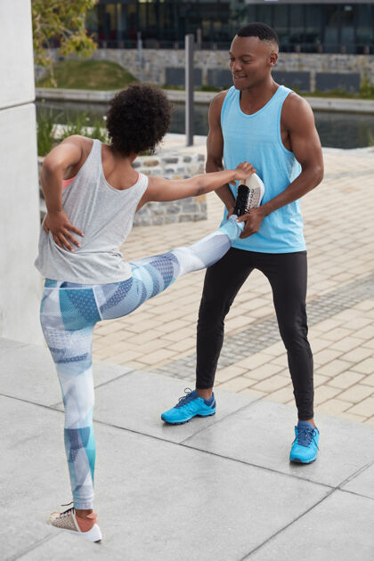 运动年轻黑皮肤女人的垂直镜头有背痛 抬腿 和教练一起做伸展运动 在外面摆姿势团结 运动 训练的概念黑人帮助锻炼女子种族垂直