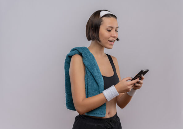 人一位身穿运动服 肩上扛着毛巾的年轻健身女士站在白墙上 面带微笑地看着手机屏幕健身站市民