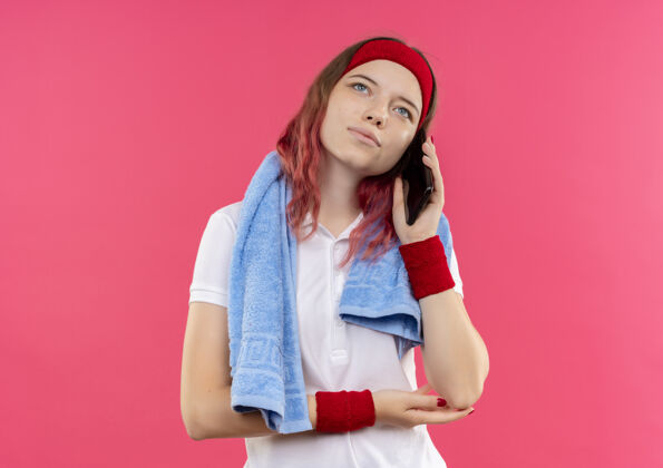 看戴着头巾 肩上扛着毛巾 面带梦幻般微笑的年轻运动女性站在粉色的墙上 一边用手机交谈运动年轻女性