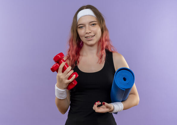 哑铃身穿运动服的年轻健身女士 手拿瑜伽垫和哑铃 站在紫色的墙上 脸上洋溢着幸福的笑容抱着快乐运动