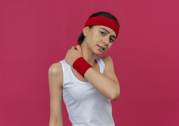 市民穿着运动服的年轻健身女士戴着头带 看起来不舒服 摸着脖子站在粉红色的墙上疼站疼痛运动员