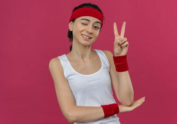 人身穿运动服的年轻健身女 头上戴着眼圈 微笑着 粉红色的墙上立着胜利的标志站立表情微笑