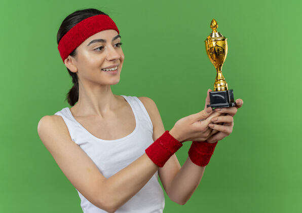 人身穿运动服 头箍 手持奖杯 面带微笑 自信地站在绿色墙壁上的年轻健身女士运动装健康运动