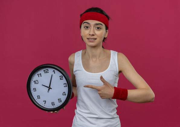 拿着穿着运动服的年轻健身女 戴着头巾 拿着挂钟 手指指着挂钟 站在粉红色的墙上 看上去很困惑看运动装女性
