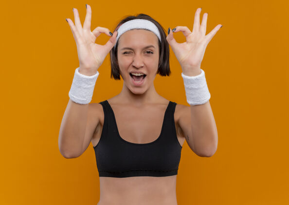 两者身穿运动服的年轻健身女 戴着头巾 微笑着眨眼 双手站在橙色的墙上做着“ok”的手势姿势人手
