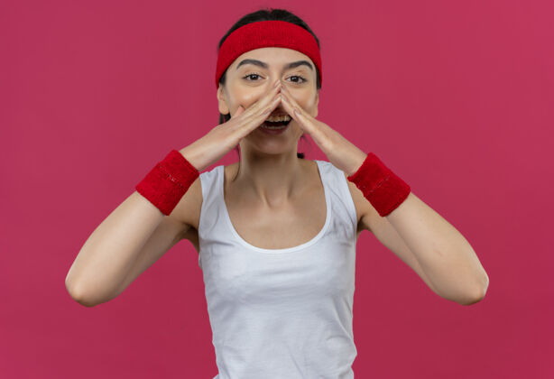 人身穿运动服的年轻健身女 戴着头巾 站在粉红色的墙上 双手靠近嘴边大声喊叫健身近姿势