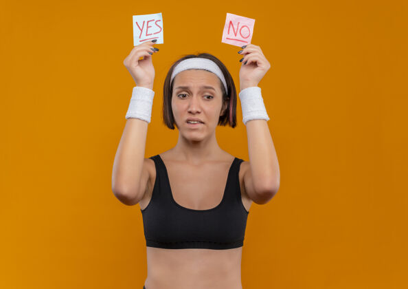 姿势身穿运动服 头箍的年轻健身女士举着两张写着“是”和“否”的提醒纸 站在橙色的墙上 神情困惑头带举起是