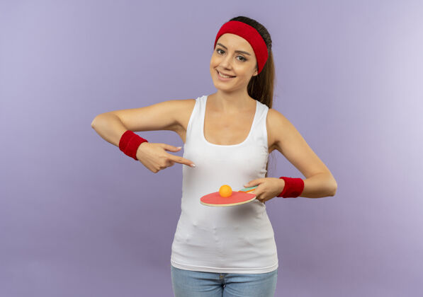 目录身穿运动服的年轻健身女士 戴着头巾 手里拿着球拍和乒乓球 用食指指着球拍 脸上带着微笑站在灰色的墙上运动女人站着